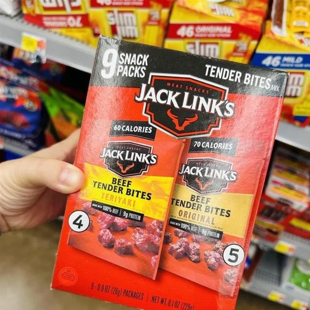 Jack Link's Beef Jerky snack pack box featuring 9 packs of tender bites in Teriyaki and Original flavors, held in a retail store aisle with blurred shelves in the background.