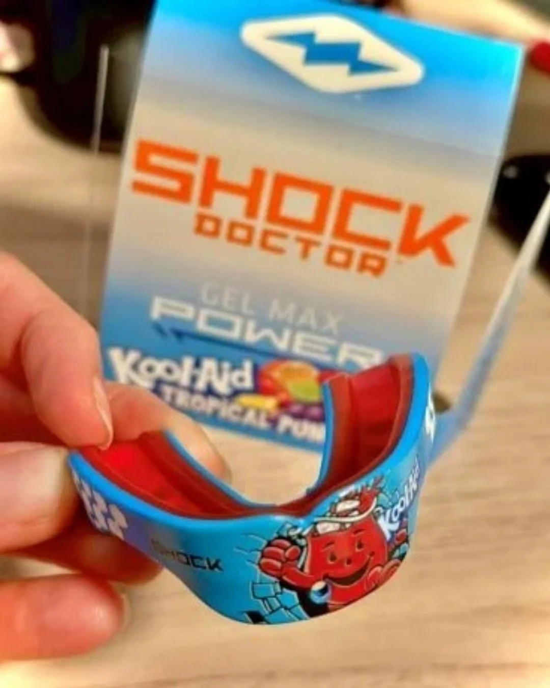 Hand holding a blue and red Shock Doctor Gel Max Power mouthguard featuring Kool-Aid branding with the Kool-Aid Man graphic, against a background showing the product packaging with Tropical Punch theme.