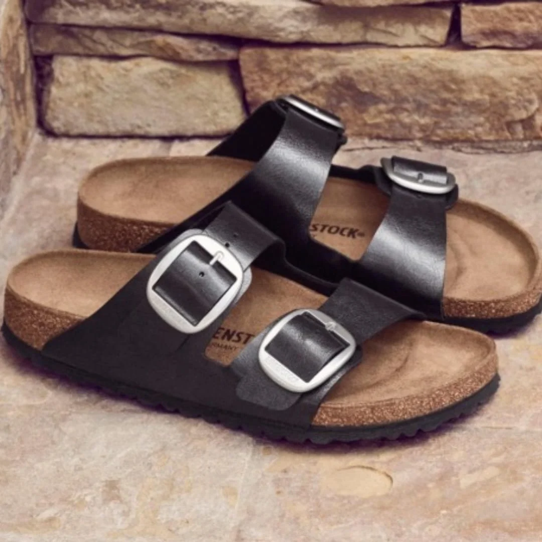 Pair of black Birkenstock Arizona Big Buckle sandals with oversized silver buckles, contoured cork footbeds, and black soles, displayed on a light stone surface with a rustic stone wall backdrop.
