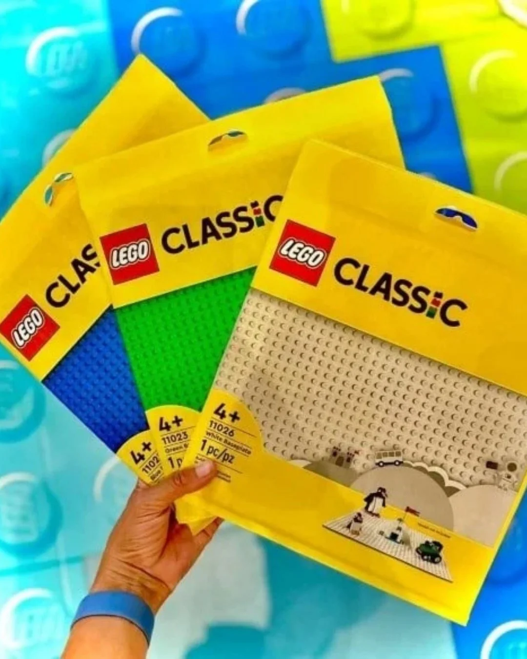 Hand holding three LEGO Classic baseplates in yellow packaging: white, green, and blue, with LEGO branding.