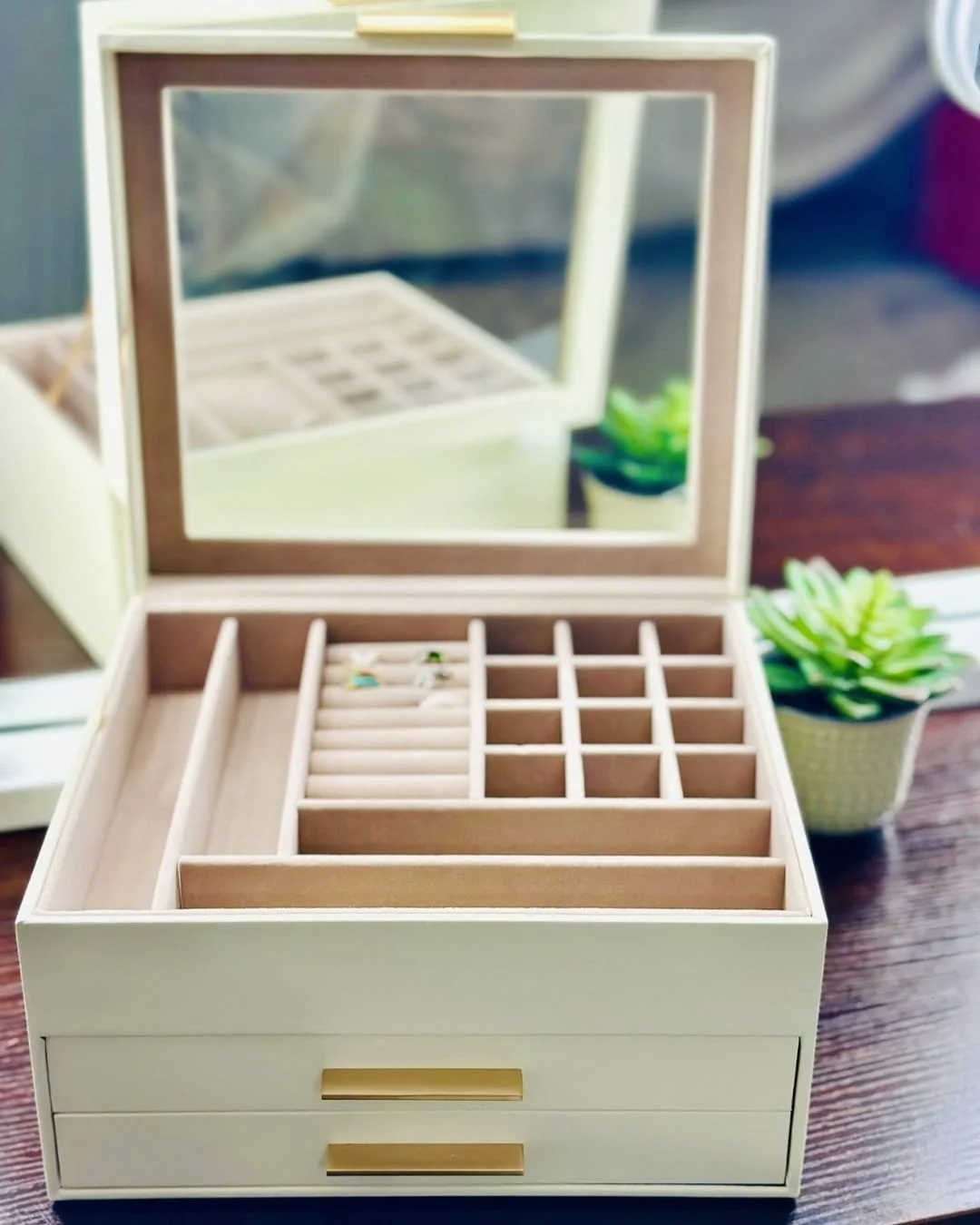 Cream jewelry box organizer with a glass lid, ring slots, divided compartments, and two drawers with gold handles, displayed on a dark wood surface beside small potted plants.