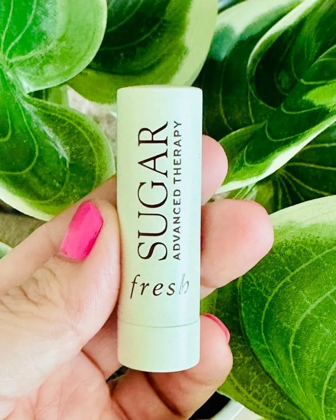 Hand holding fresh Sugar Advanced Therapy Lip Treatment tube against green leaves