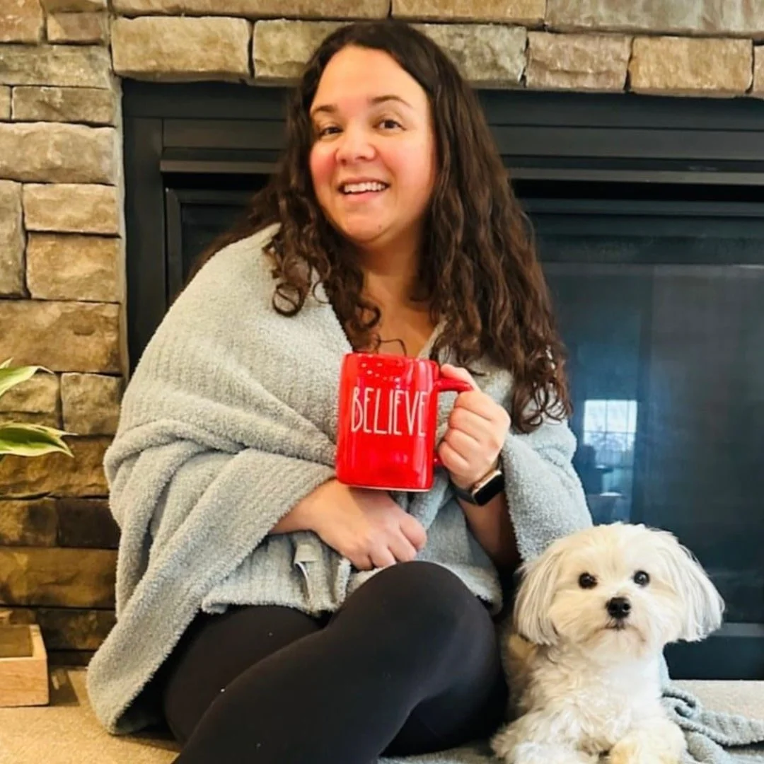 Nicole, CEO of Nicole's Fabulous Finds, smiling while holding a red BELIEVE mug, seated next to a small white dog in front of a stone fireplace.