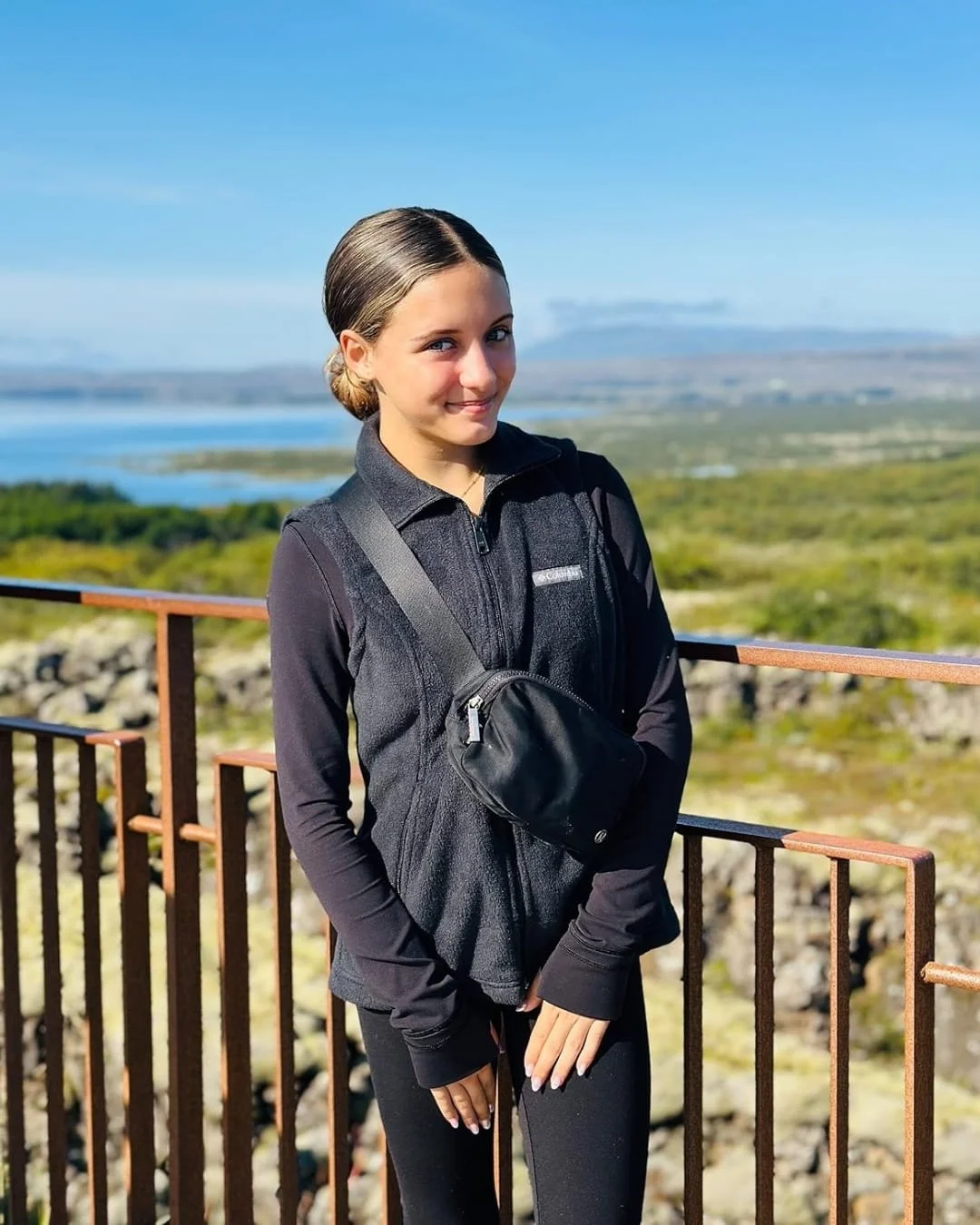 Black cross-body belt bag worn by a young woman standing behind a metal railing with a scenic backdrop of a lake, greenery, and distant mountains under a clear blue sky.