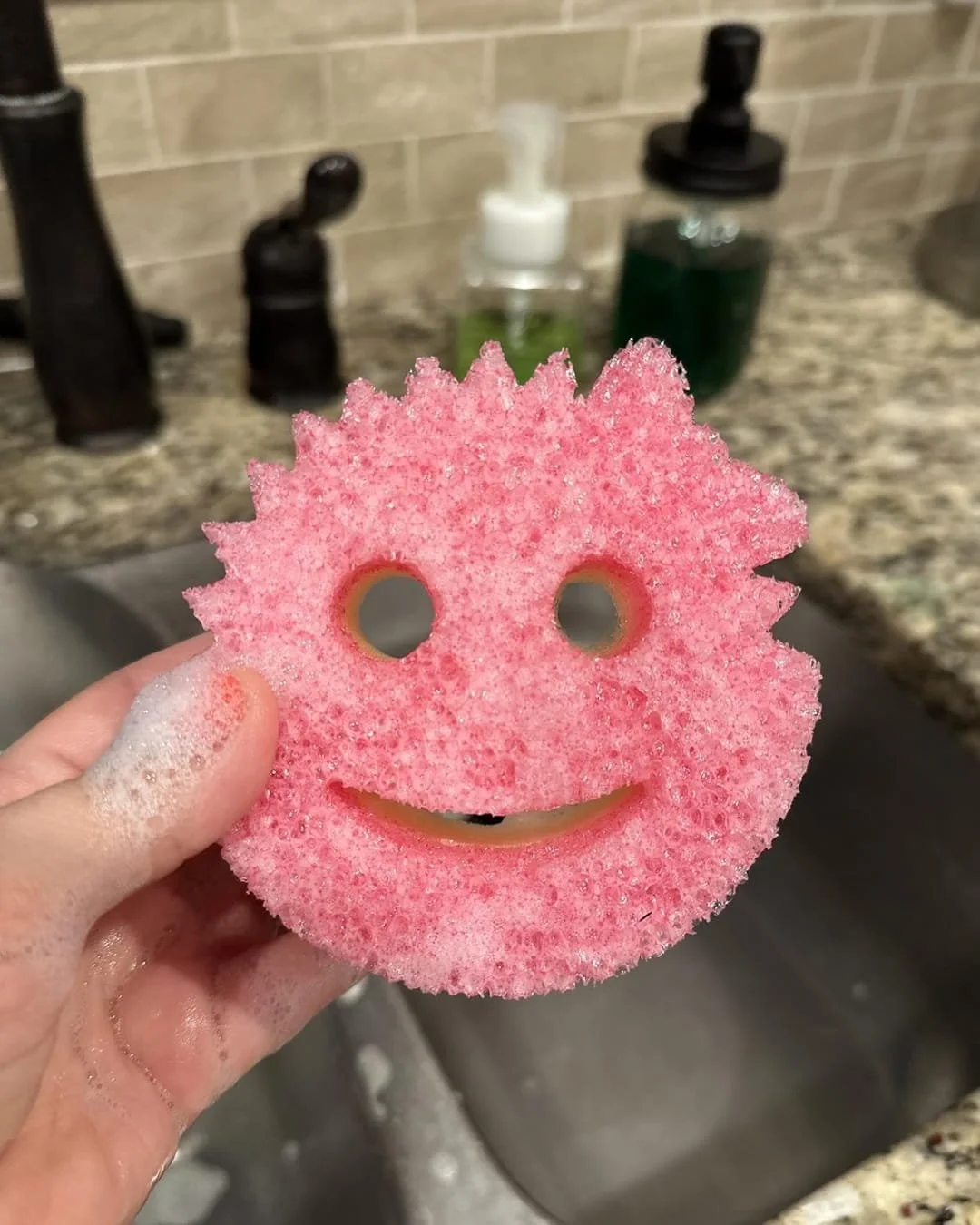 Pink Scrub Mommy sponge with a smiling face design, held by a soapy hand over a modern kitchen sink with soap dispensers and a granite countertop in the background.