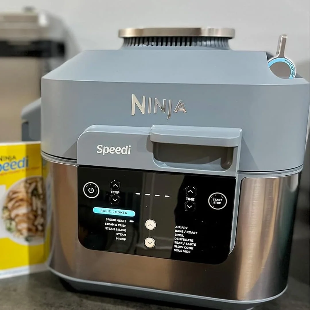 Ninja Speedi Rapid Cooker and Air Fryer with a dual-tone blue-grey and stainless steel design, featuring a detailed digital control panel and mode selection lever, displayed on a kitchen countertop with a recipe booklet in the background.
