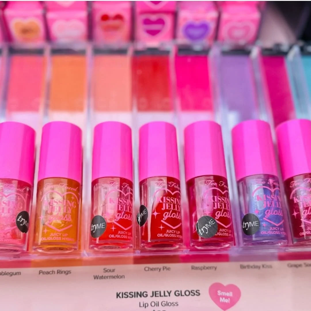 Retail display of Too Faced Kissing Jelly Lip Oil Gloss featuring seven vibrant shades with pink caps, candy-inspired names like 'Peach Rings' and 'Sour Watermelon,' and heart-themed branding.
