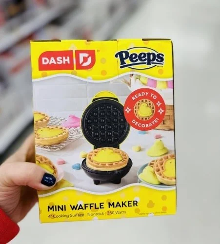 Box for Dash Peeps Mini Waffle Maker, featuring a yellow design with images of waffles topped with Peeps chicks, held by a hand with blue-painted nails.