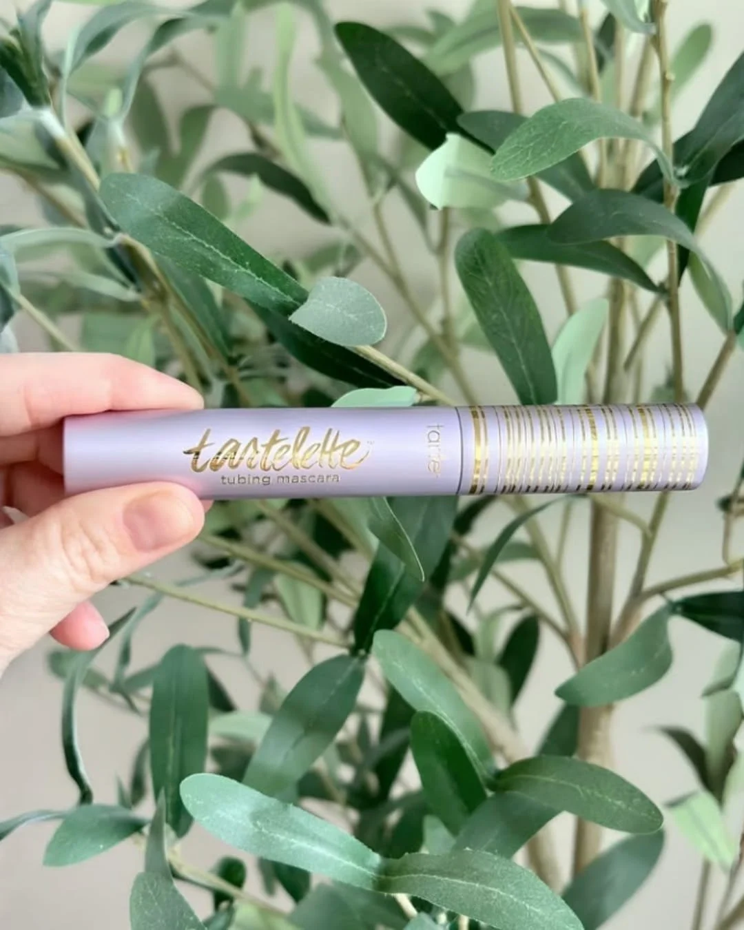 Lavender Tartelette Tubing Mascara tube with gold accents held in front of a leafy green background