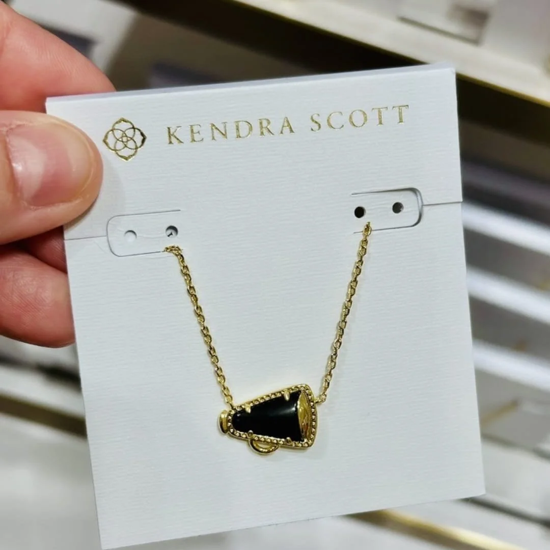 Kendra Scott necklace featuring a gold-toned megaphone pendant with a black inlay, displayed on a branded white jewelry card held by a hand.