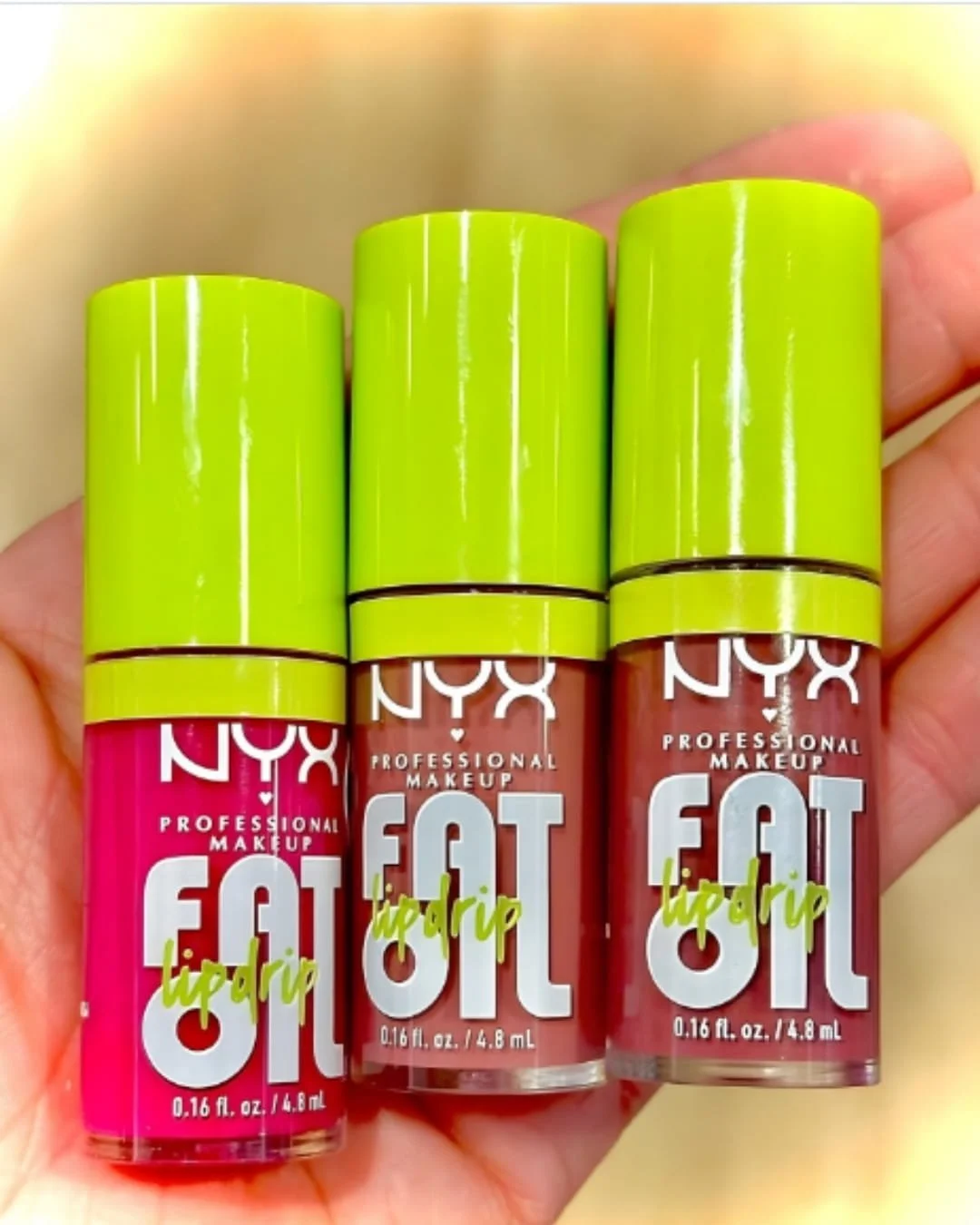 Three NYX Professional Makeup Fat Oil Lip Drip glosses in neon pink, terracotta, and mauve shades, featuring bold white and neon green typography, with chunky lime-green caps, held in a hand against a soft yellow background.