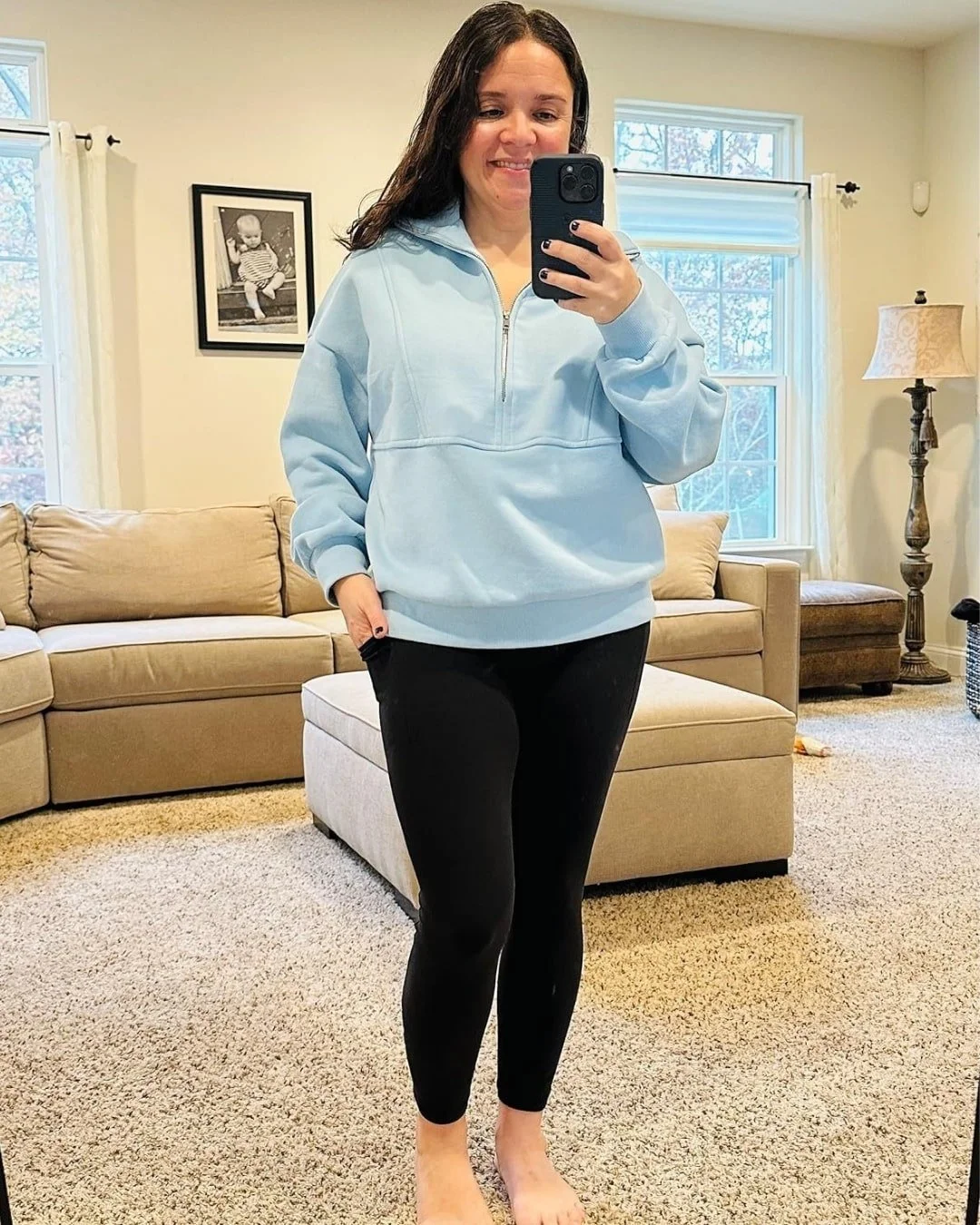 Nicole from NicFab Finds wearing a cozy light blue quarter-zip sweater and black leggings, taking a mirror selfie in her warm, inviting living room.