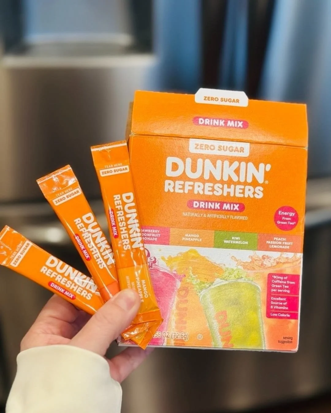 Dunkin' Refreshers Zero Sugar Drink Mix variety pack featuring flavors like Mango Pineapple and Strawberry Dragonfruit Lemonade, with individual stick packets held in a hand against a kitchen background.