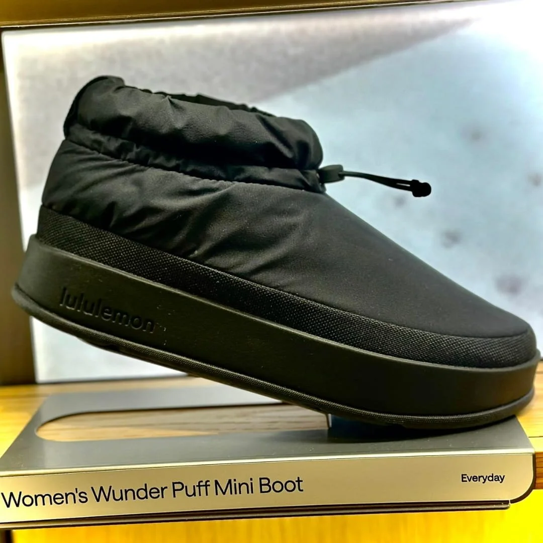Lululemon Women's Wunder Puff Mini Boot in black with a quilted upper, drawstring closure, and thick sole featuring embossed branding, displayed on a metallic retail stand.