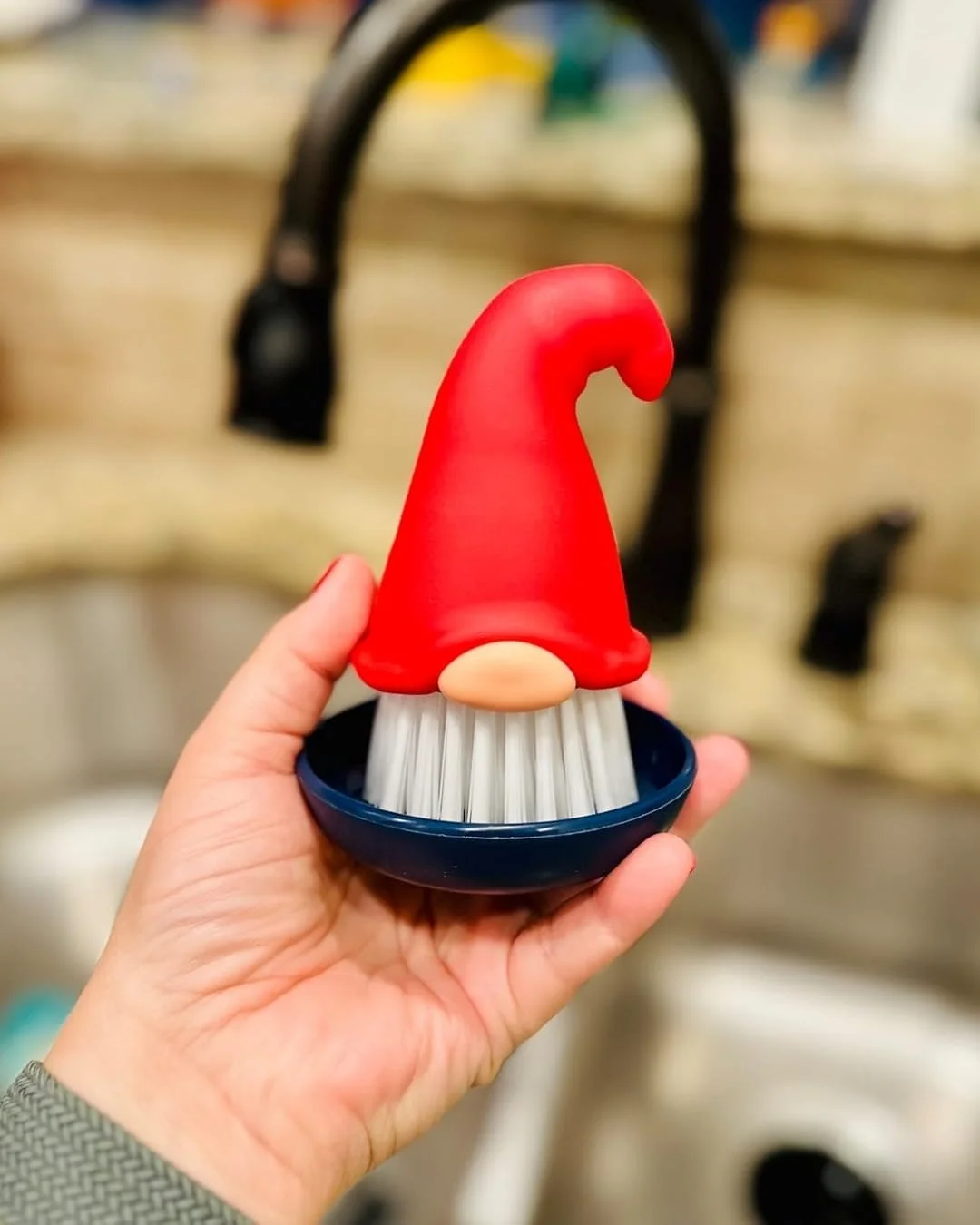 Whimsical Beardy Gnome dish brush with a red hat handle, white bristle beard, and blue base, held by a hand in a kitchen setting with a blurred faucet and countertop in the background.