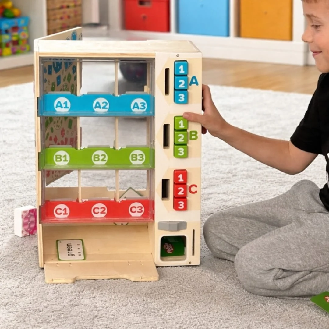 Child playing with a wooden vending machine toy featuring a color-coded alphanumeric grid system (A1-A3 in blue, B1-B3 in green, C1-C3 in red), interactive buttons, and a dispensing slot, set in a bright playroom with a gray shag carpet.