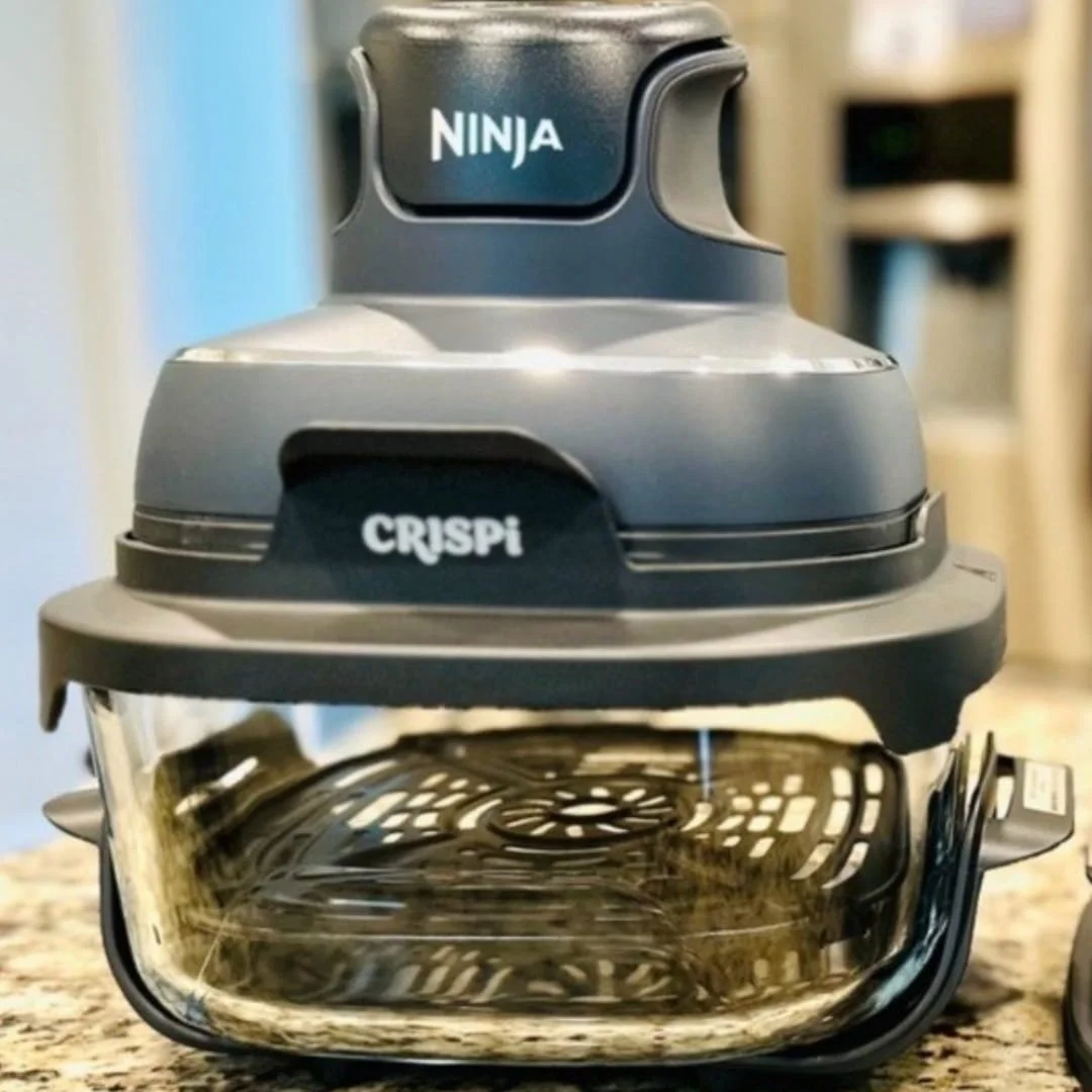 Ninja Crispi air fryer with a sleek dark grey design, featuring a clear glass cooking chamber with a black crisper tray inside, placed on a granite kitchen countertop with blurred cabinetry in the background.