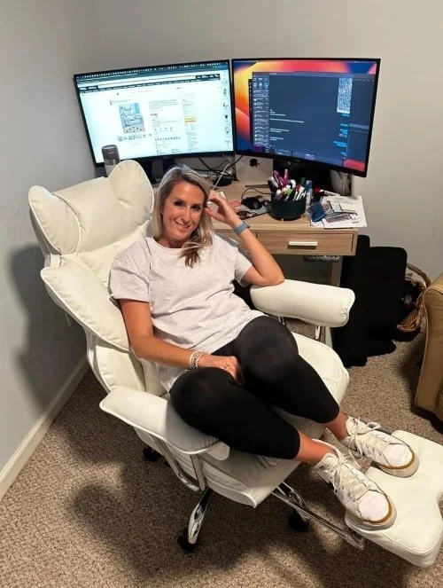Woman sitting in a white reclining office chair with a footrest, smiling at the camera in a home office setup with a desk, dual monitors displaying an e-commerce site and a dark interface, and light neutral decor.