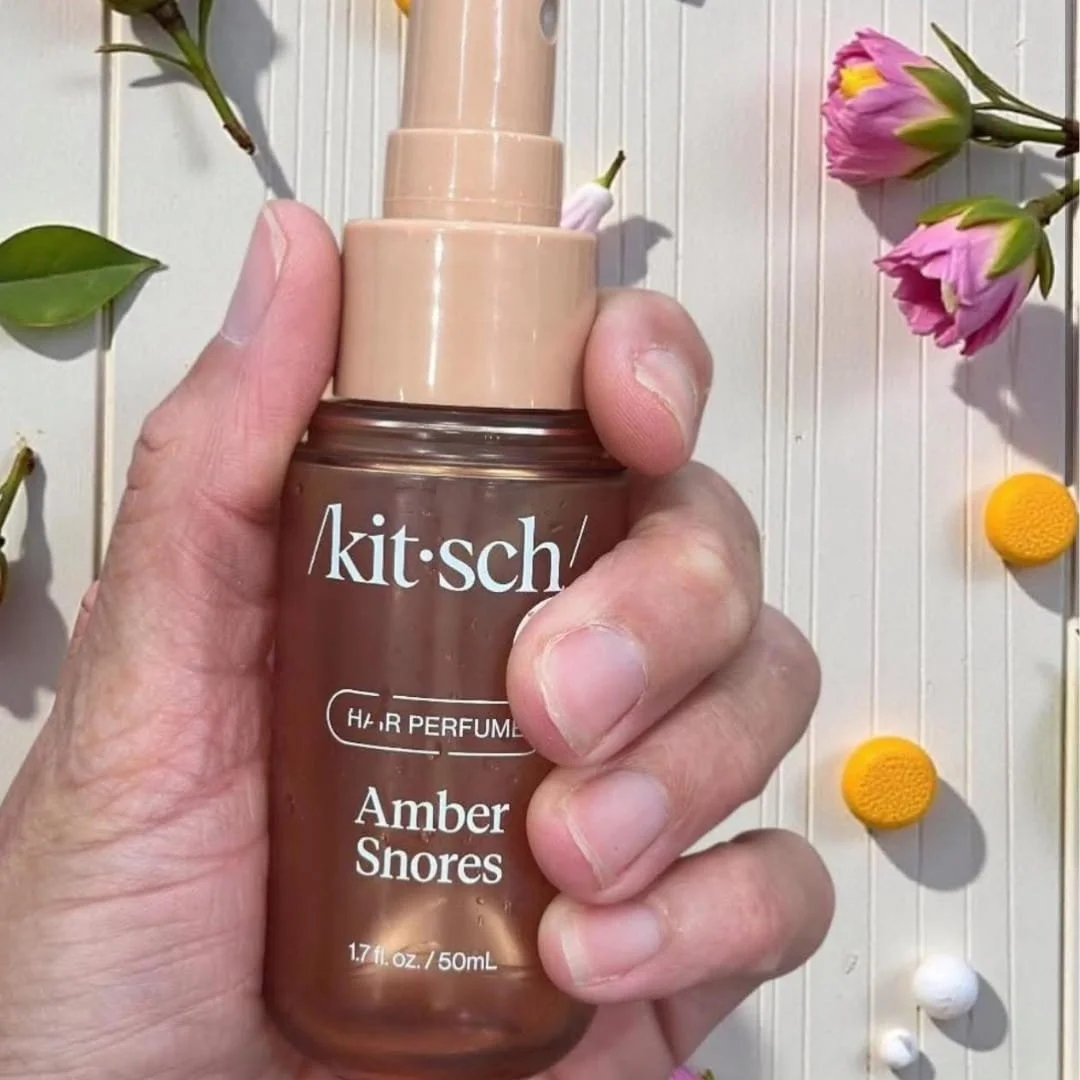 Kitsch Amber Hair Perfume bottle with amber-colored liquid, held by a hand against a white wooden background, surrounded by flowers and natural decorative elements.