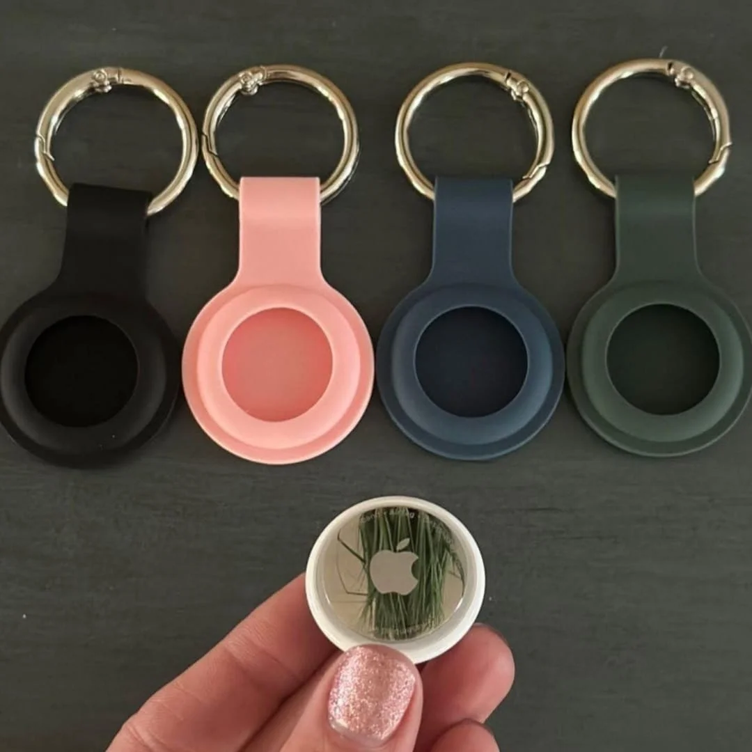 Apple AirTag with colorful silicone keychain holders in black, pink, blue, and green, featuring a sleek metallic design and Apple logo, showcased on a dark textured background.