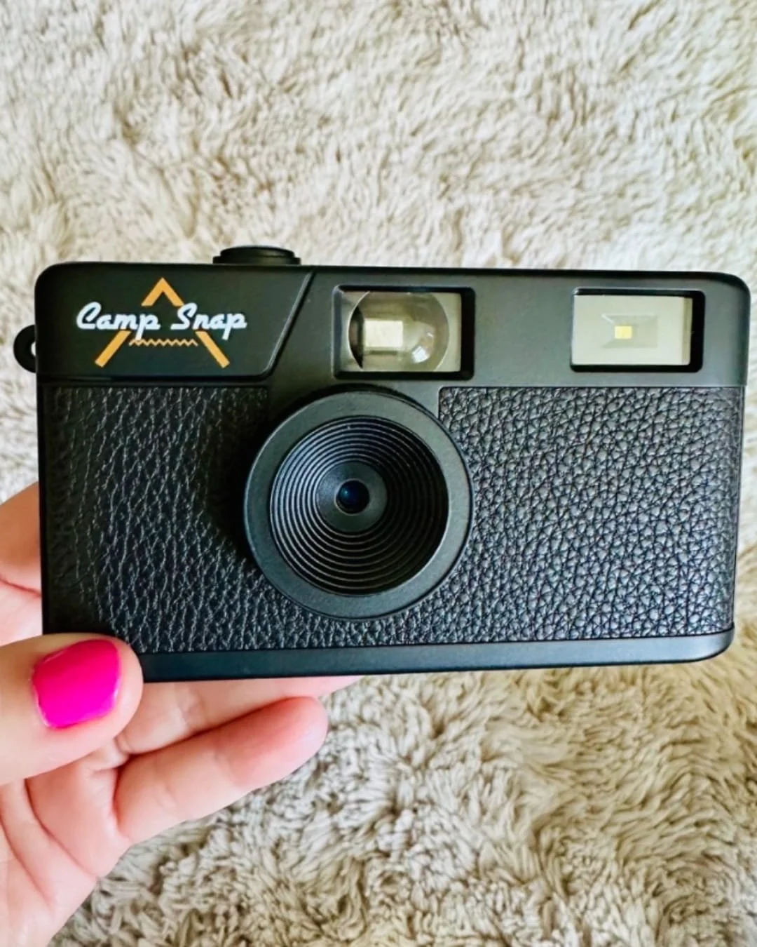 Camp Snap screenless digital camera with a retro black design, featuring a faux-leather texture, yellow branding, and a vintage aesthetic, held by a hand with bright pink nail polish against a beige textured background.