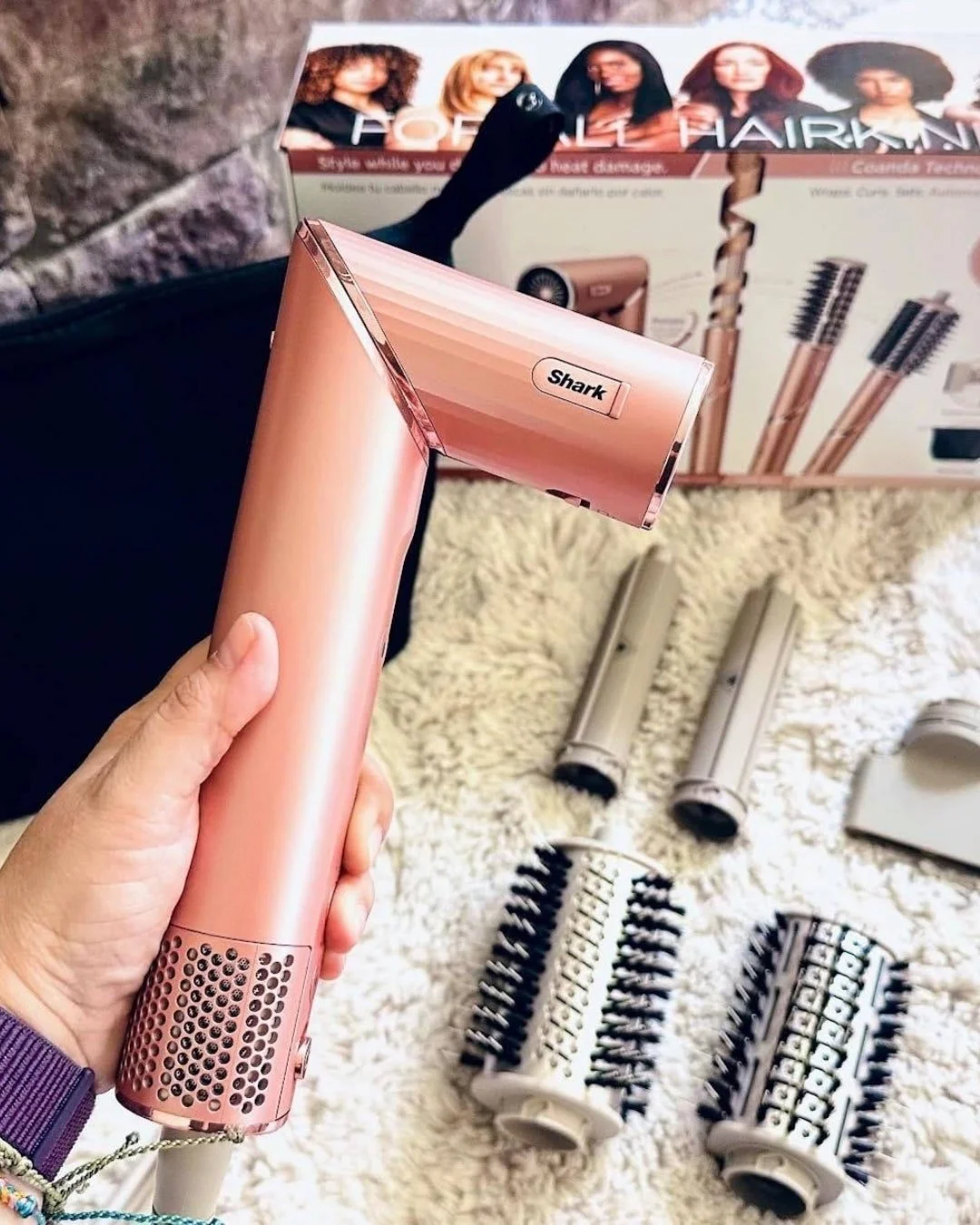 Rose gold Shark hair styling device with multiple attachments, including curling barrels and brushes, displayed on a fluffy white rug with product packaging in the background highlighting features for all hair types and heat damage protection.