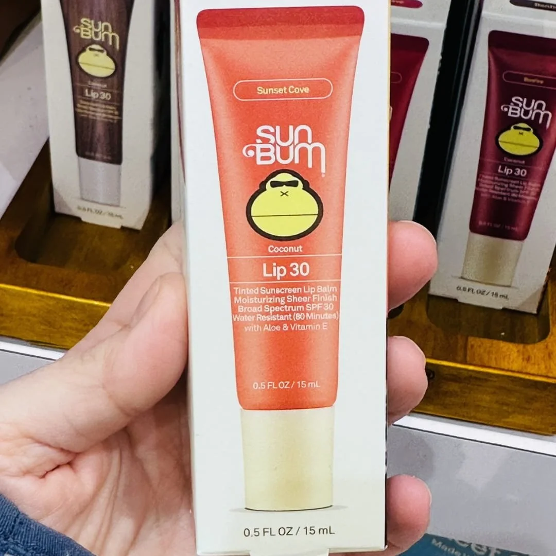 Sun Bum Lip 30 Tinted Sunscreen Lip Balm in Sunset Cove flavor, featuring coral packaging with SPF 30 protection, held in a hand against a wooden retail display with other balm varieties.