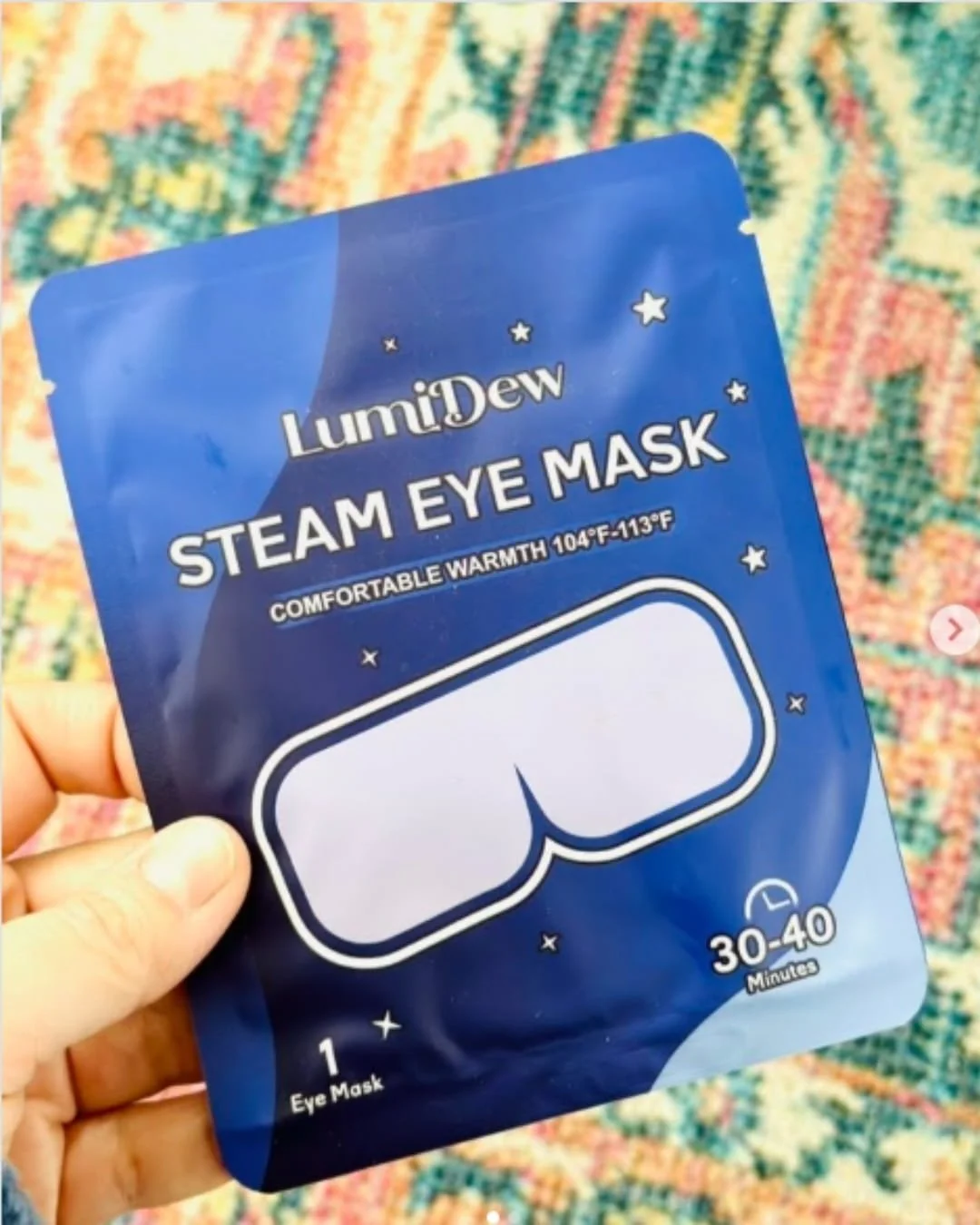 Hand holding a LumiDew Steam Eye Mask packet in dark blue packaging with a mask illustration, warmth details, and star accents against a colorful patterned background