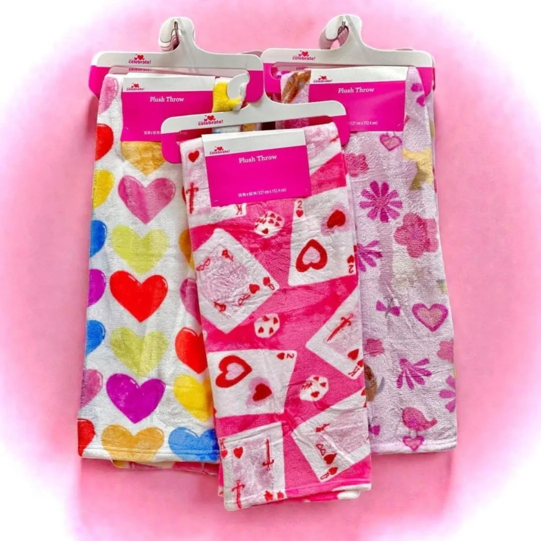 Celebrate! Valentine's Day plush throw blankets in vibrant patterns, including colorful hearts, playing card designs, and floral motifs, displayed on white hangers against a pink background.