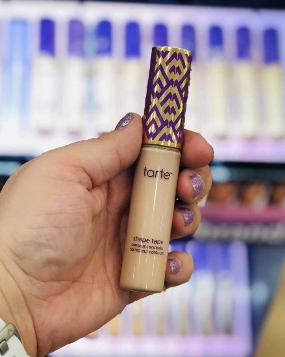 Tarte Shape Tape creamy concealer tube held in hand with makeup store display in background