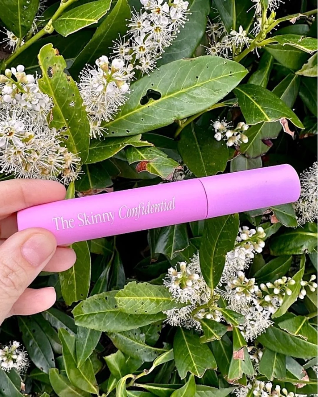 The Skinny Confidential Brow Peptide in a bright pink cylindrical tube, held horizontally by a hand with a lush green Cherry Laurel plant and white flowers in the background, showcasing a vibrant and natural aesthetic.
