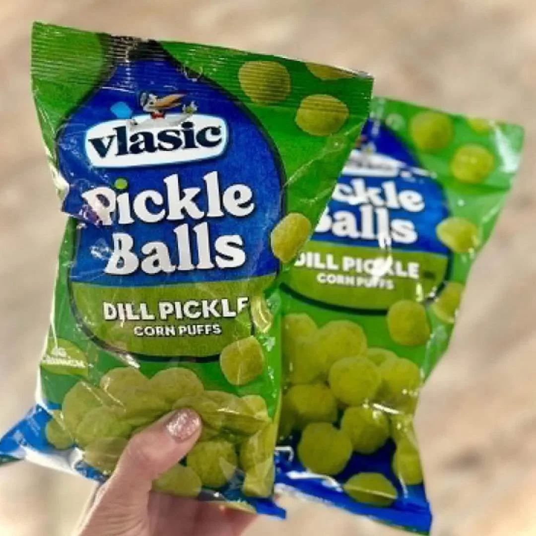 Two bags of Vlasic Pickle Balls Dill Pickle Corn Puffs, featuring green and blue packaging with the Vlasic logo, held by a hand with glittery nails against a light background.
