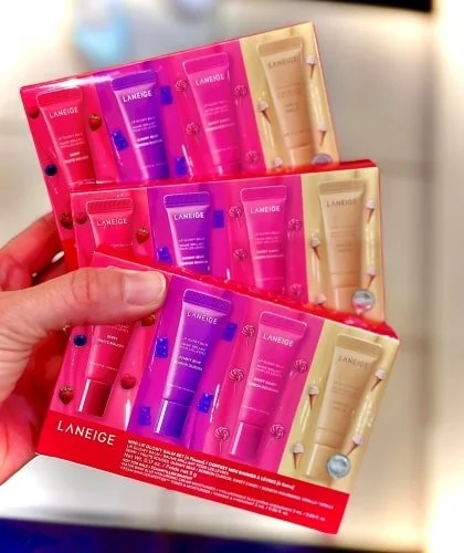 Hand holding a LANEIGE Lip Glowy Balm gift set featuring three flavors: Berry, Gummy Bear, and Sweet Candy, in vibrant packaging with thematic illustrations, stacked with additional sets in the background.