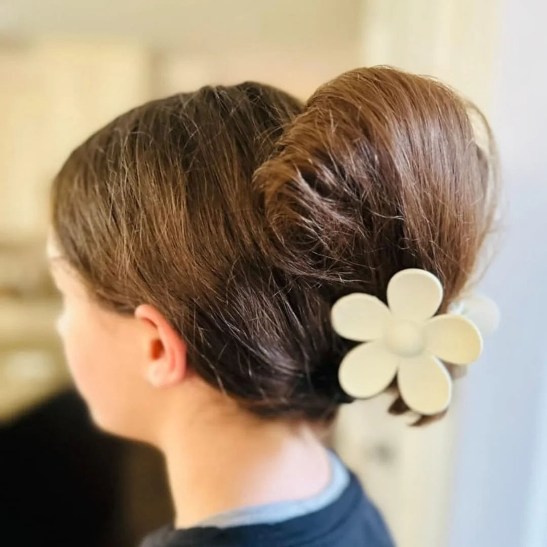 Back view of a messy bun hairstyle secured with a cream-colored flower-shaped hair claw clip, worn by a person with brown hair in a softly lit indoor setting.