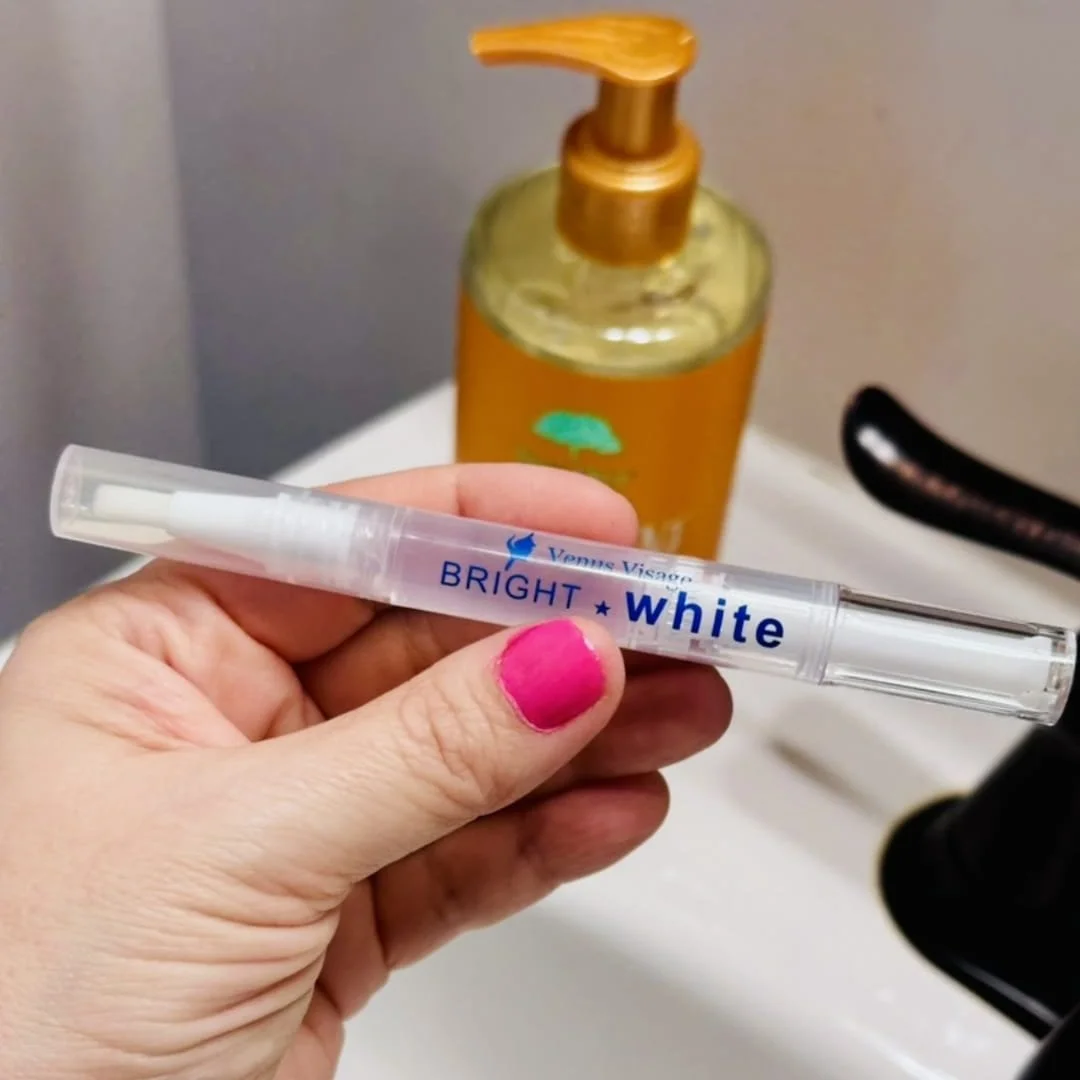 Venus Visage Bright White teeth-whitening pen with a clear gel-filled body and white brush tip, held by a hand with pink nails over a bathroom sink, with a soap bottle and faucet in the blurred background.