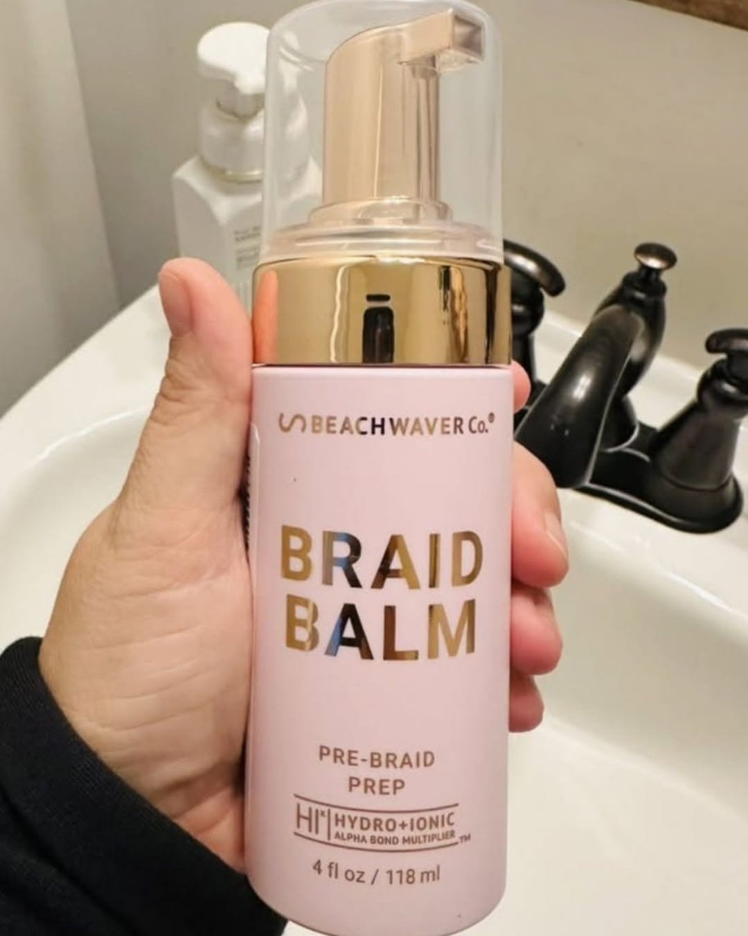 Hand holding Beachwaver Primer Foam in a pink bottle with gold pump, photographed over a bathroom sink