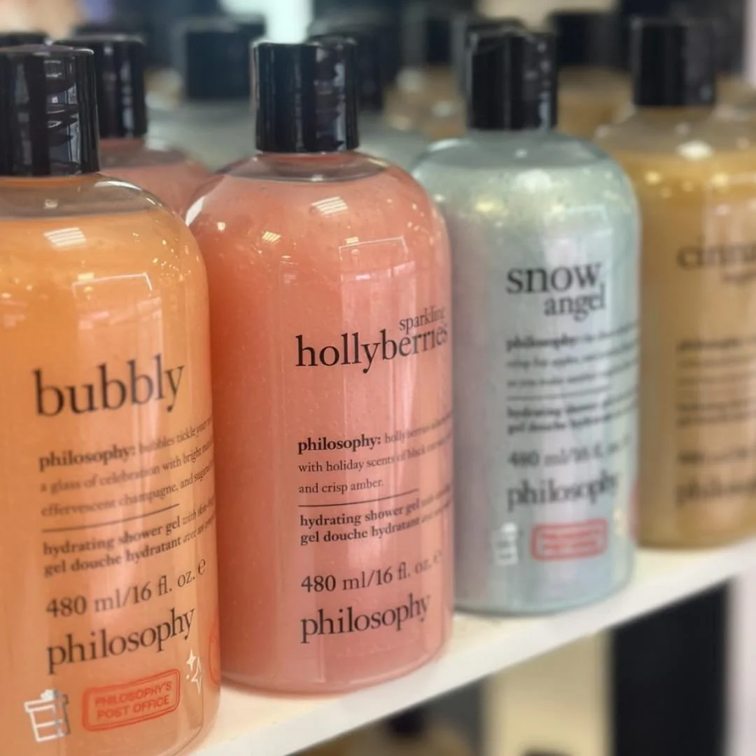 Philosophy hydrating shower gels in festive scents including Bubbly, Sparkling Hollyberries, Snow Angel, and Cinnabun, displayed on a white shelf in a retail setting.