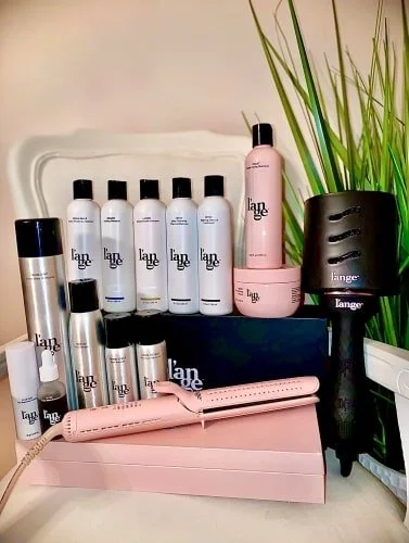 Collection of Lange hair care products and styling tools, including white bottles, rose gold flat iron, and a black diffuser, arranged on a white surface with green plants in the background.