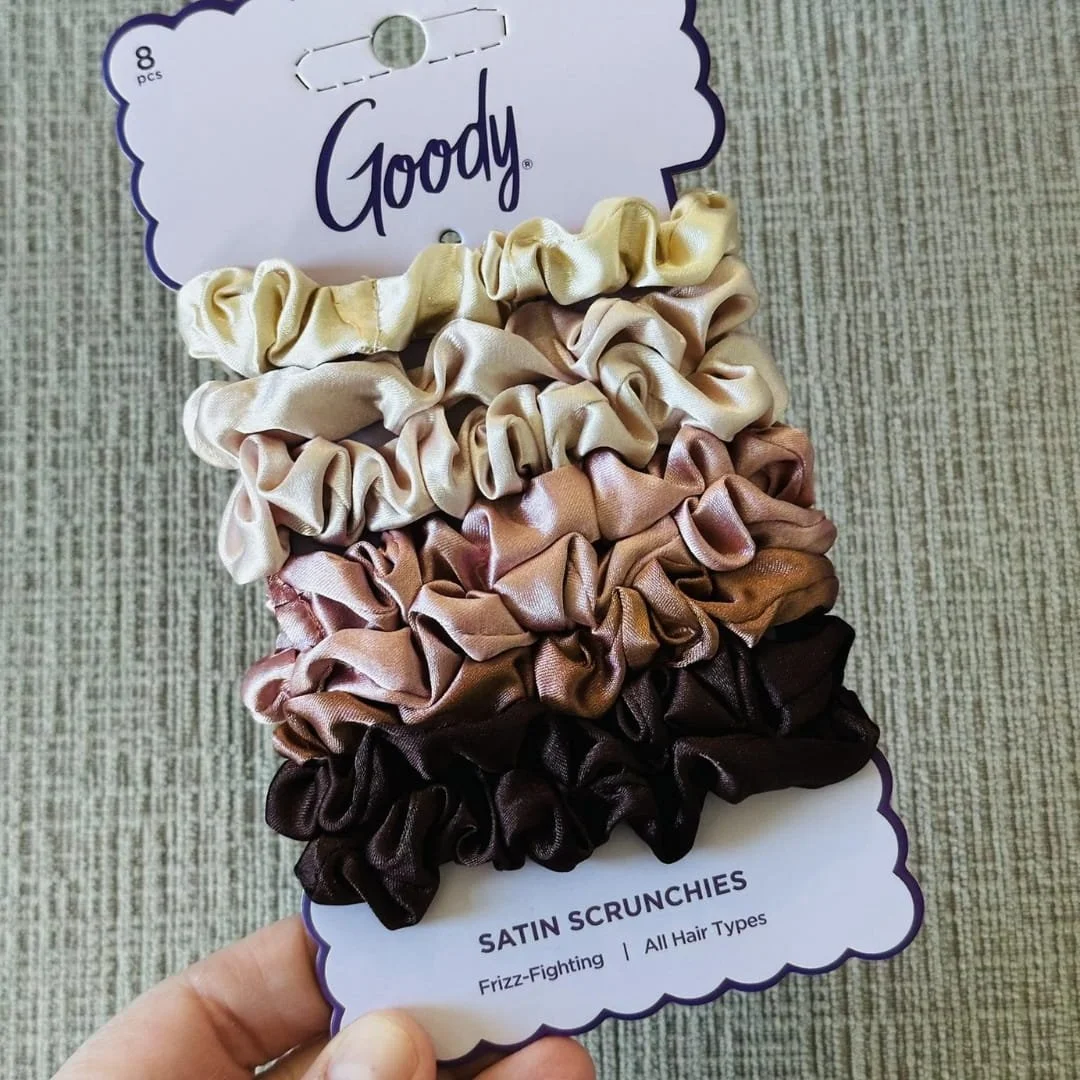 Goody Satin Scrunchies 8-pack featuring neutral tones from cream to dark brown, mounted on a white scalloped card with purple branding, held against a textured fabric background.