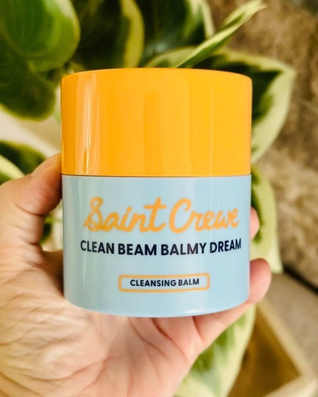 Saint Crewe Clean Beam Balmy Dream cleansing balm in a vibrant orange and light blue jar, held by a hand against a blurred green and yellow botanical background, showcasing a clean and modern aesthetic.