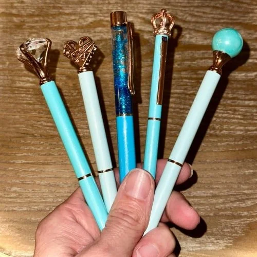 Hand holding five decorative pens in shades of blue and teal, each featuring unique metallic toppers including a crystal, heart, glitter barrel, crown, and bead, set against a wooden background.