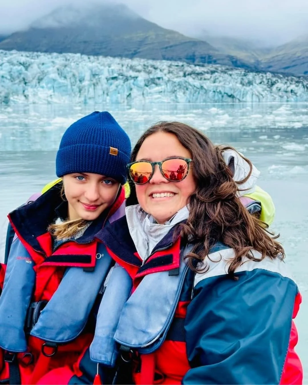 Two people wearing cold-weather gear, including a dark blue Danish Endurance beanie and red and blue life jackets, posing in front of a glacier and icy water in a cold, misty environment.