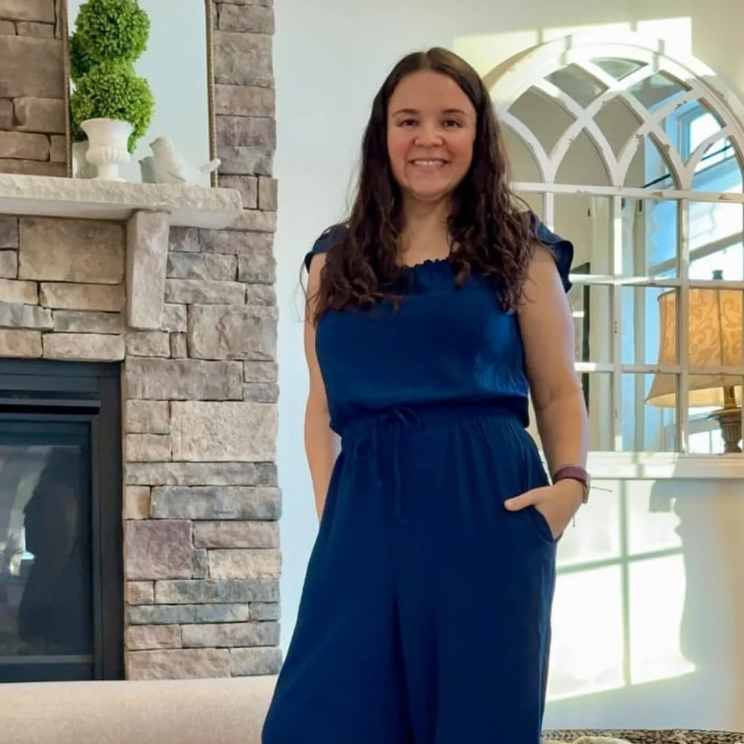 Nicole smiling indoors, wearing a dark blue jumpsuit, standing between a stone fireplace with decorative accents and an arched window frame in a well-lit room.