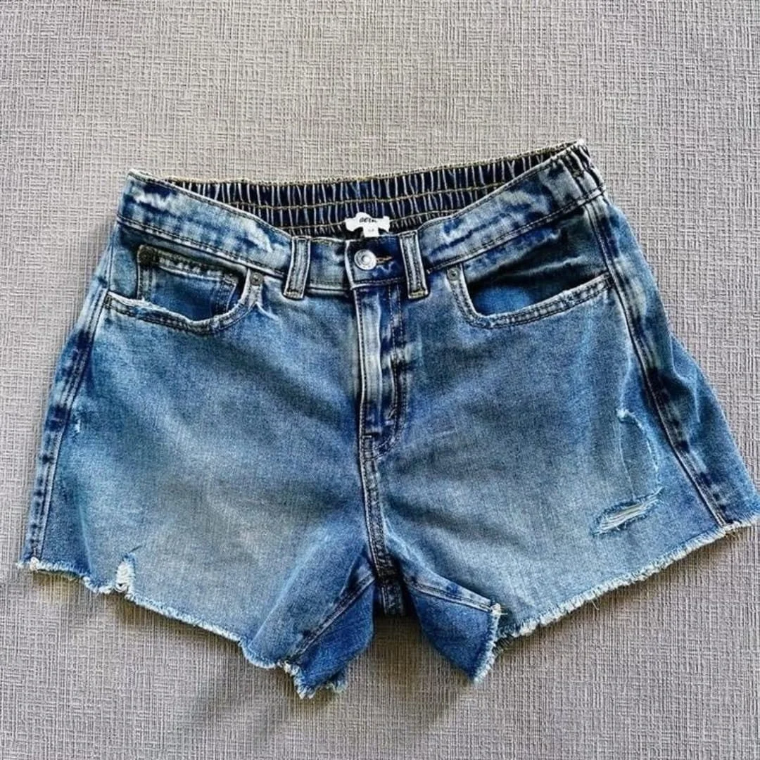 Light-wash Aerie denim shorts with a button front, elastic back waistband, distressed details, and frayed raw hem, laid flat on a textured neutral fabric background
