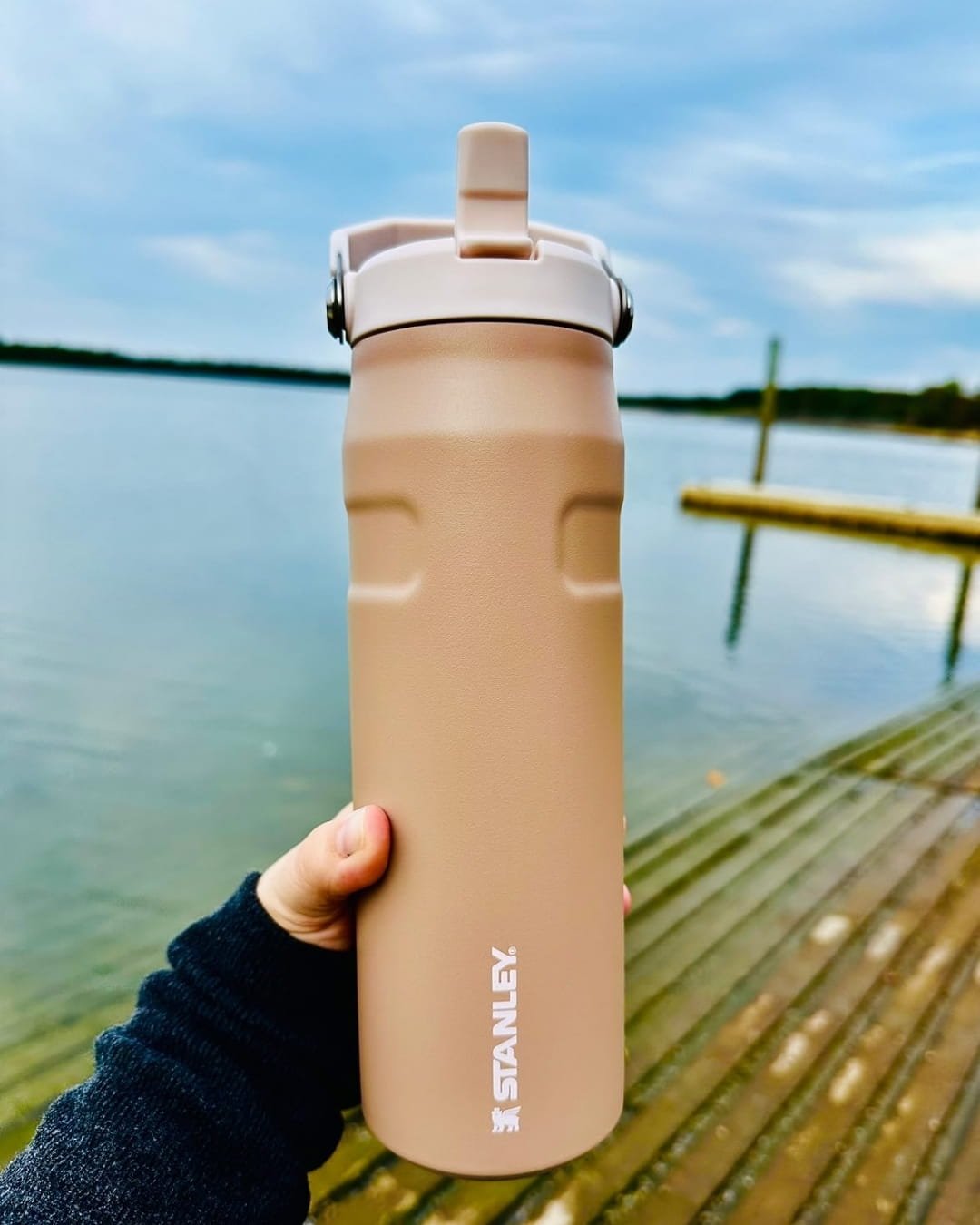 Tan Stanley Flip Straw Water Bottle with a cream lid and metal carry handle, held by a hand against a scenic backdrop of a calm lake, wooden dock, and distant shoreline under a cloudy sky.