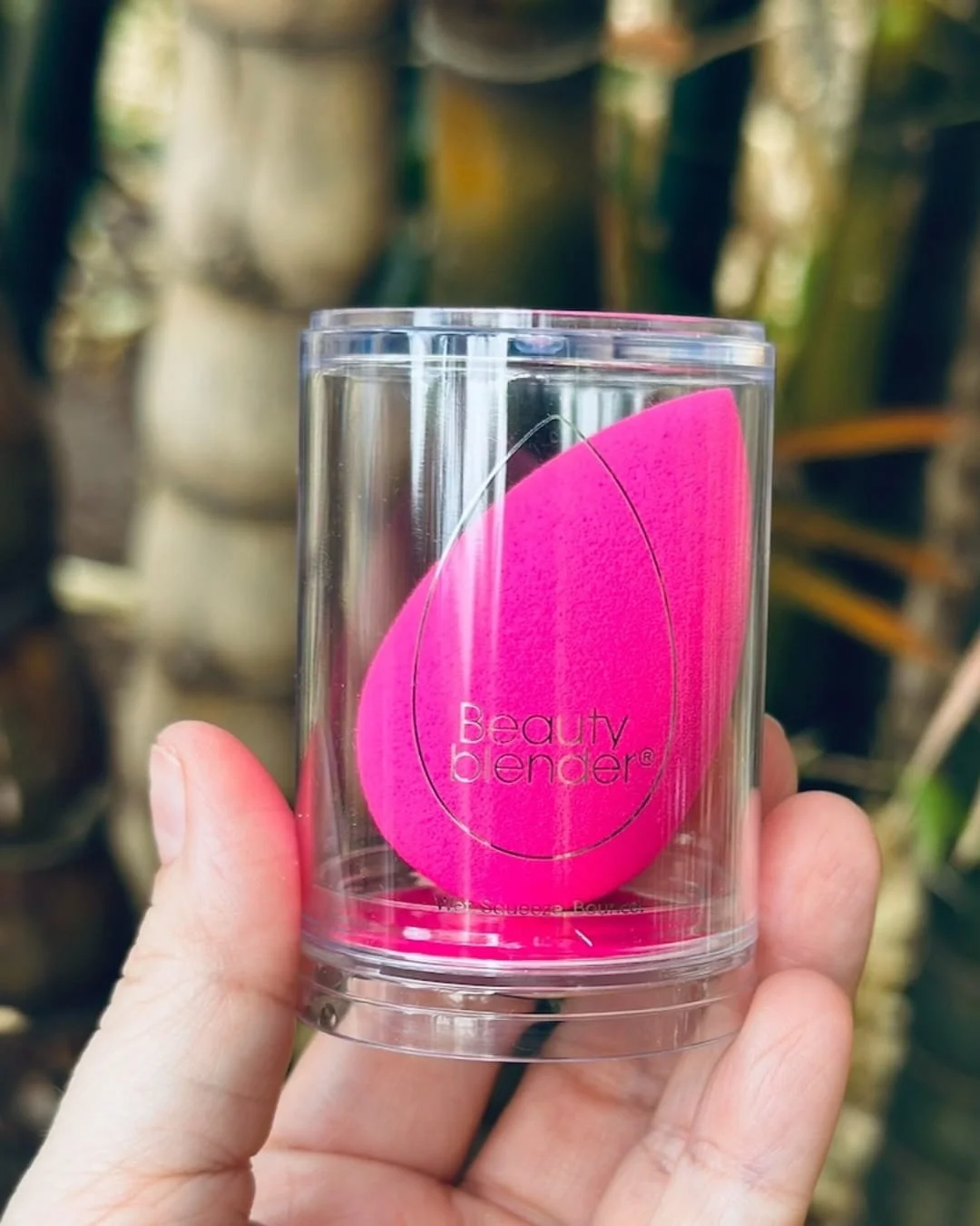 Neon pink Beautyblender makeup sponge inside a clear cylindrical container with branding, held by a hand against a blurred natural bamboo-like background.