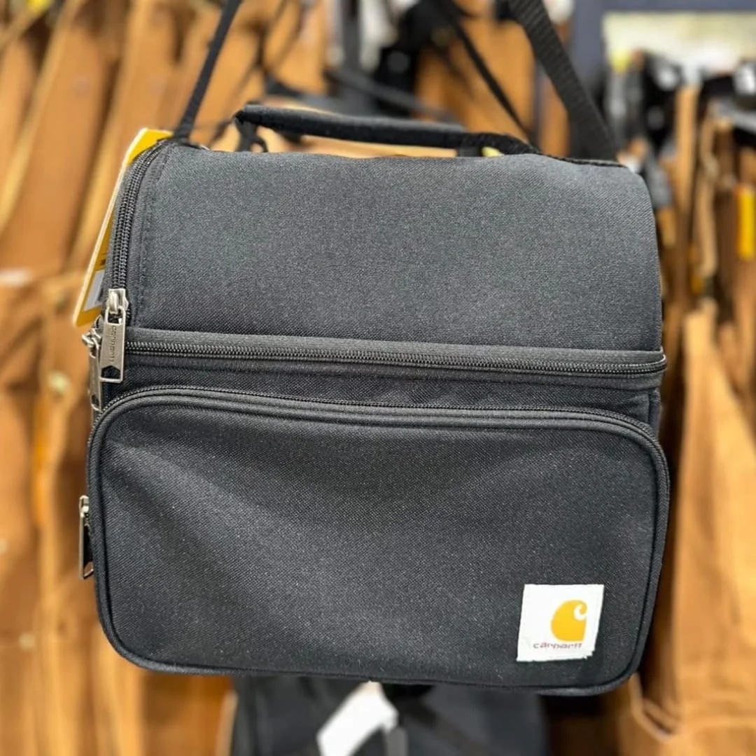 Carhartt black lunch cooler bag with dual zippered compartments, top handle, and shoulder strap, featuring the iconic Carhartt logo, displayed in a retail setting.