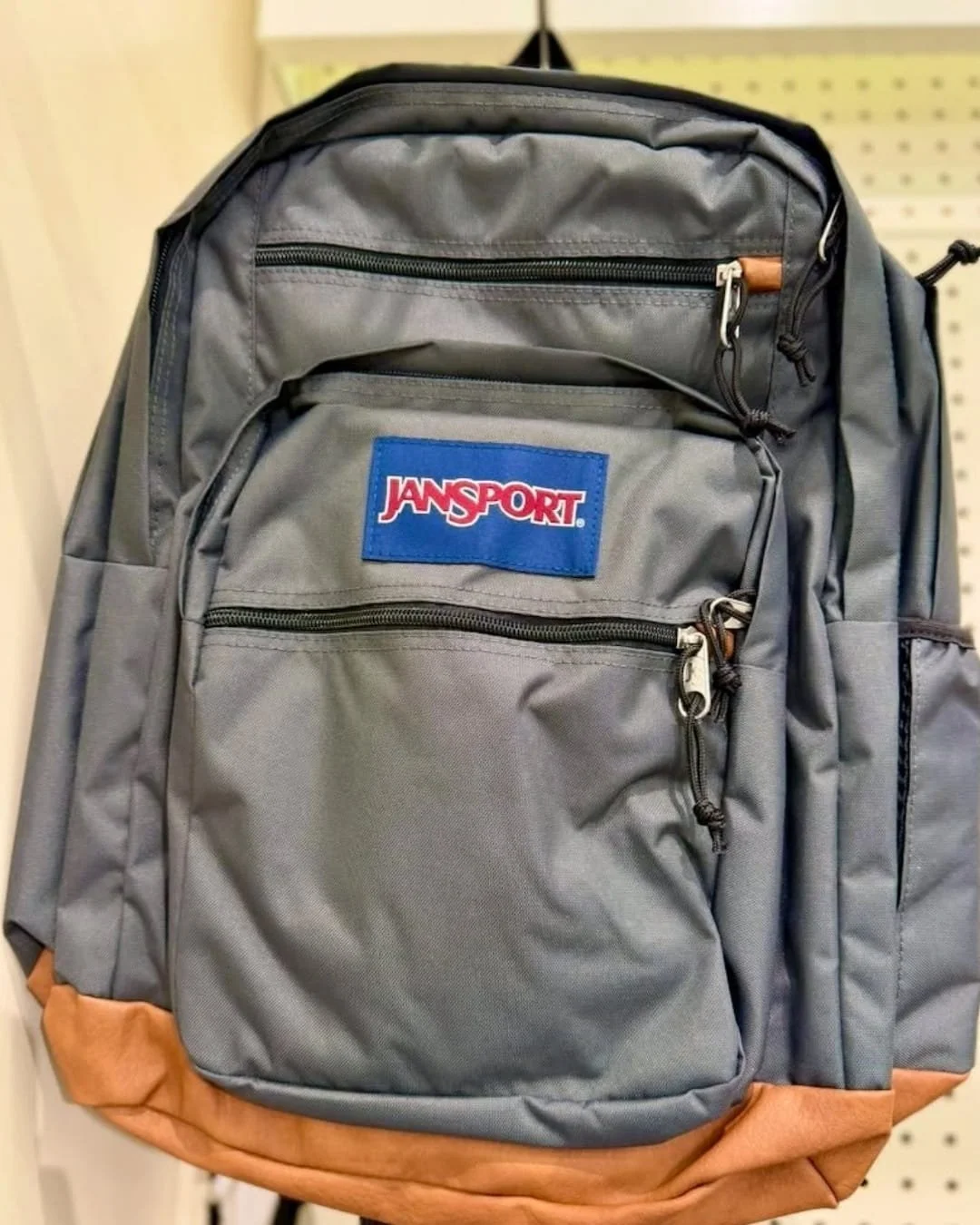 Dark gray JanSport backpack with multiple zippered compartments and a tan bottom panel displayed in a retail store