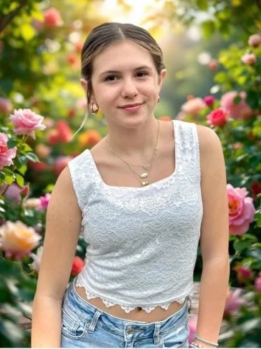 Smiling young woman in a white lace crop top and light blue jeans, standing in a vibrant rose garden with pink and red blooms, accessorized with gold jewelry.