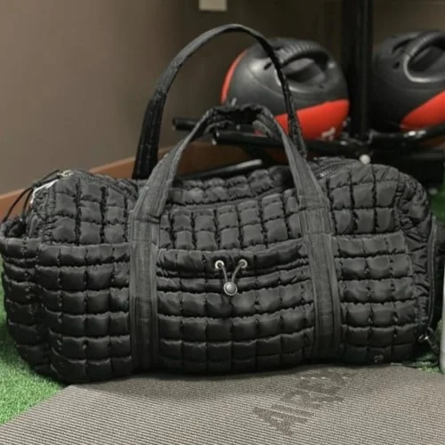 Black quilted duffel bag with padded texture and drawstring pocket, resting on a gray fitness mat, with gym equipment like medicine balls in the blurred background.